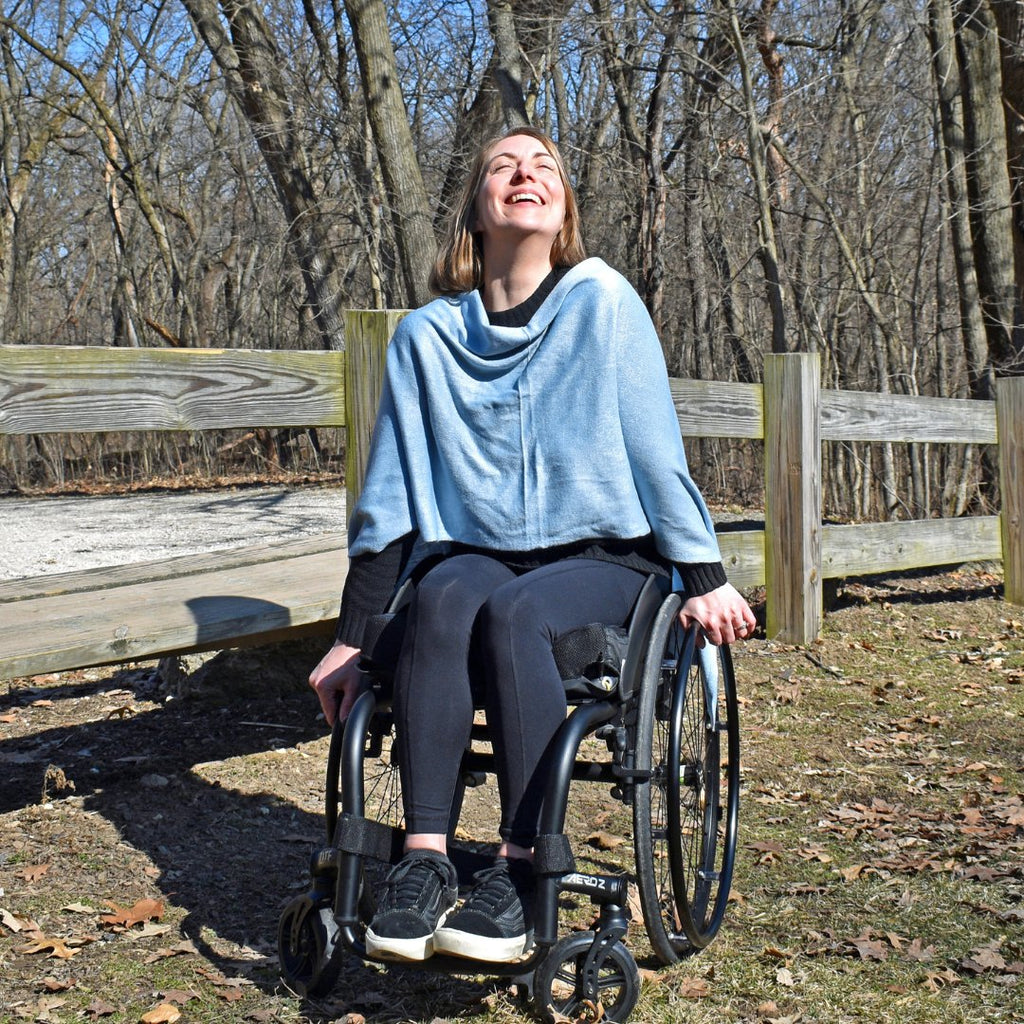 Channon, a blonde young woman is sitting in her wheelchair outside. She is wearing a blue knit wrap and is smiling.