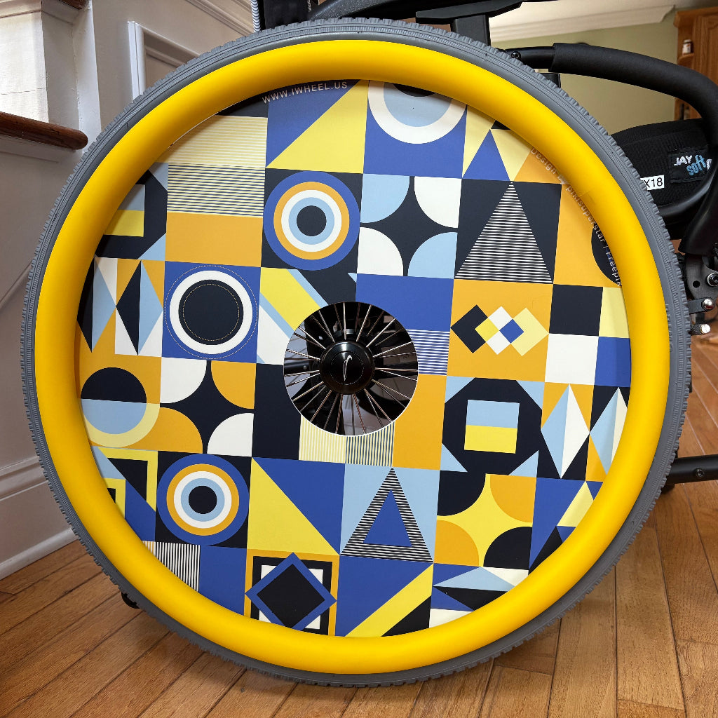 Close up picture of a rear wheelchair wheel, beautified with a vibrant yellow push rim cover and a wheel cover showing various yellow and blue geometric patterns. 