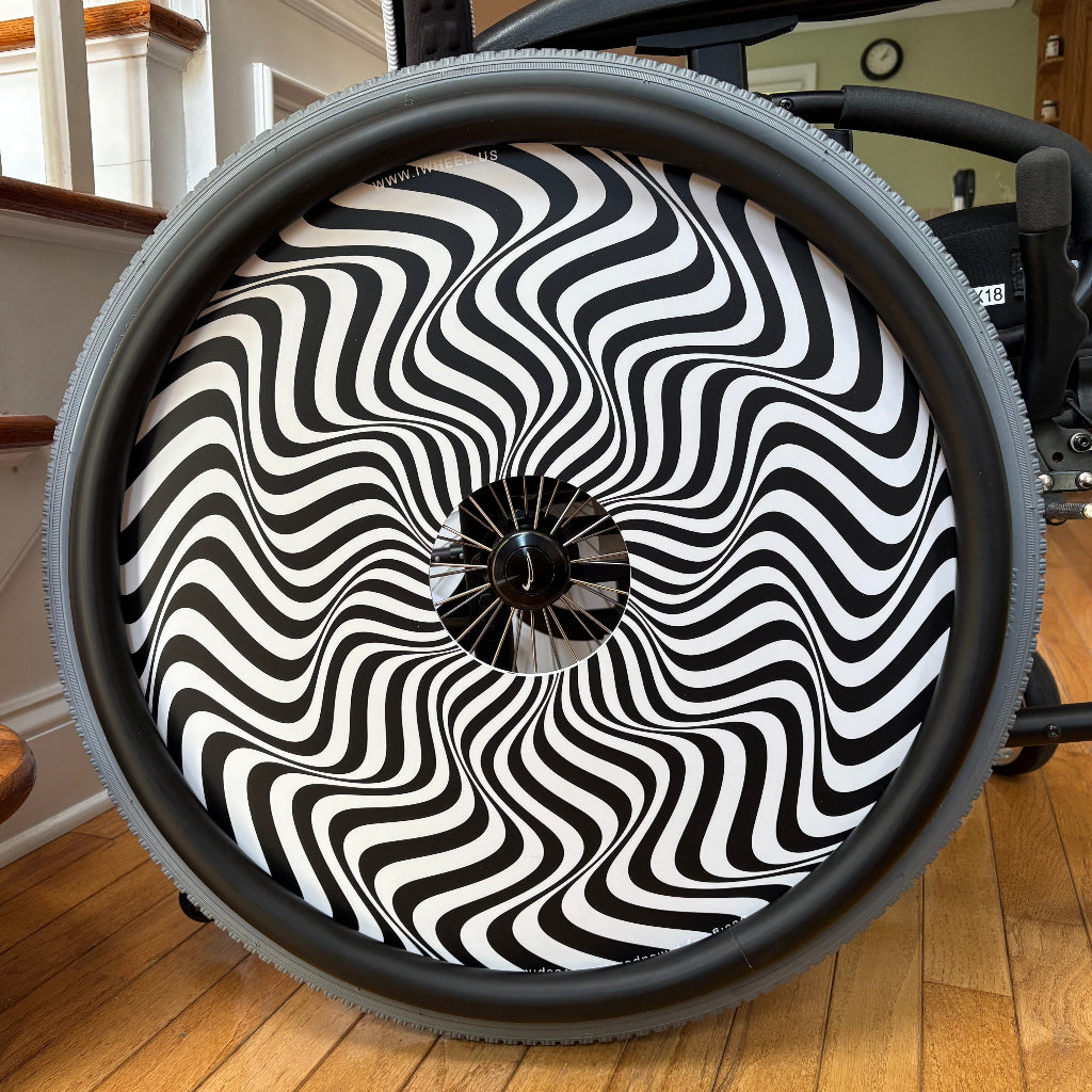 Close up picture of a wheelchair rear wheel, accessorized with a black push rim cover and a wheel cover. The wheel cover has a zig zag, black and white design. 
