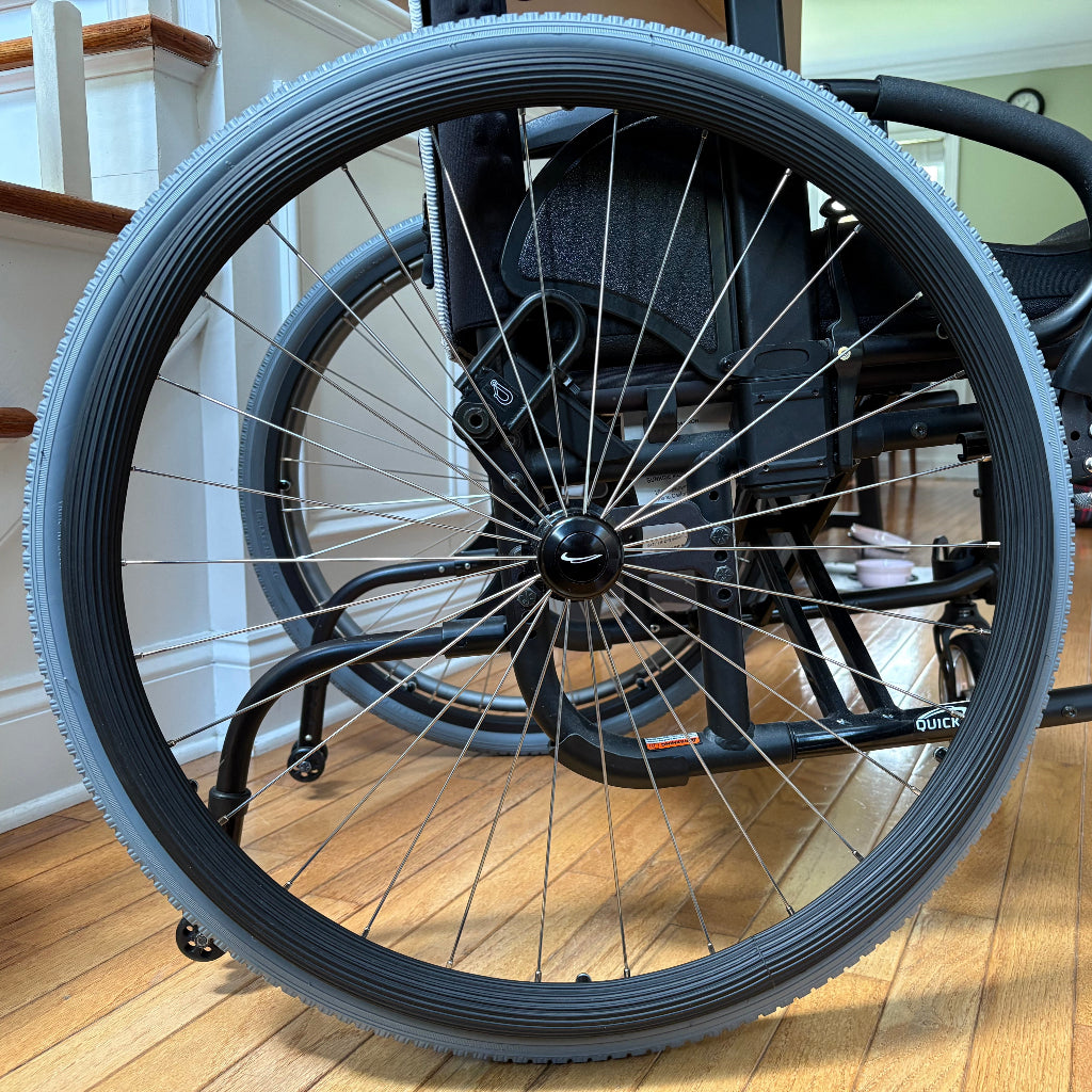 Close-up side picture of a rear wheelchair wheel, with a black grooved push rim cover on the push rim.