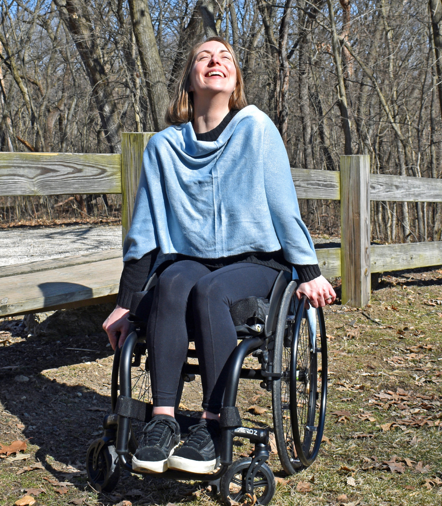 Channon is a blond woman. She is seating in her wheelchair, smiling, while enjoying a synny day outside in the park. She is wearing an adaptive, sky blue knit wrap.