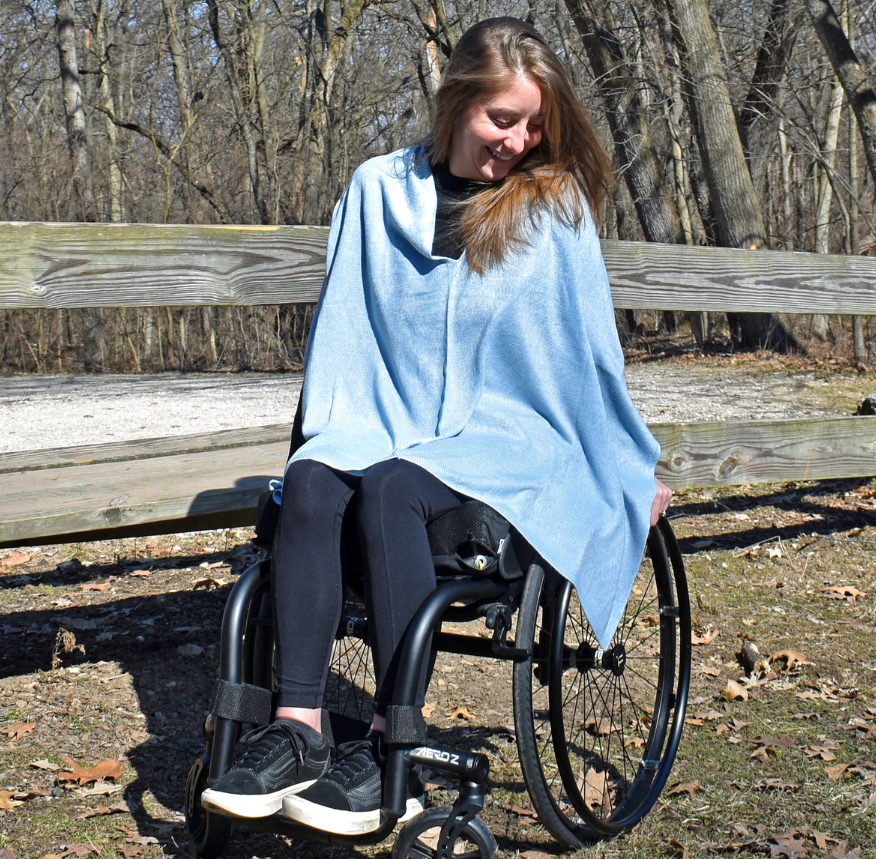 Channon is a blond woman. She is seating in her wheelchair, smiling, while enjoying a sunny day outside in the park. She is wearing an adaptive sky blue shawl.